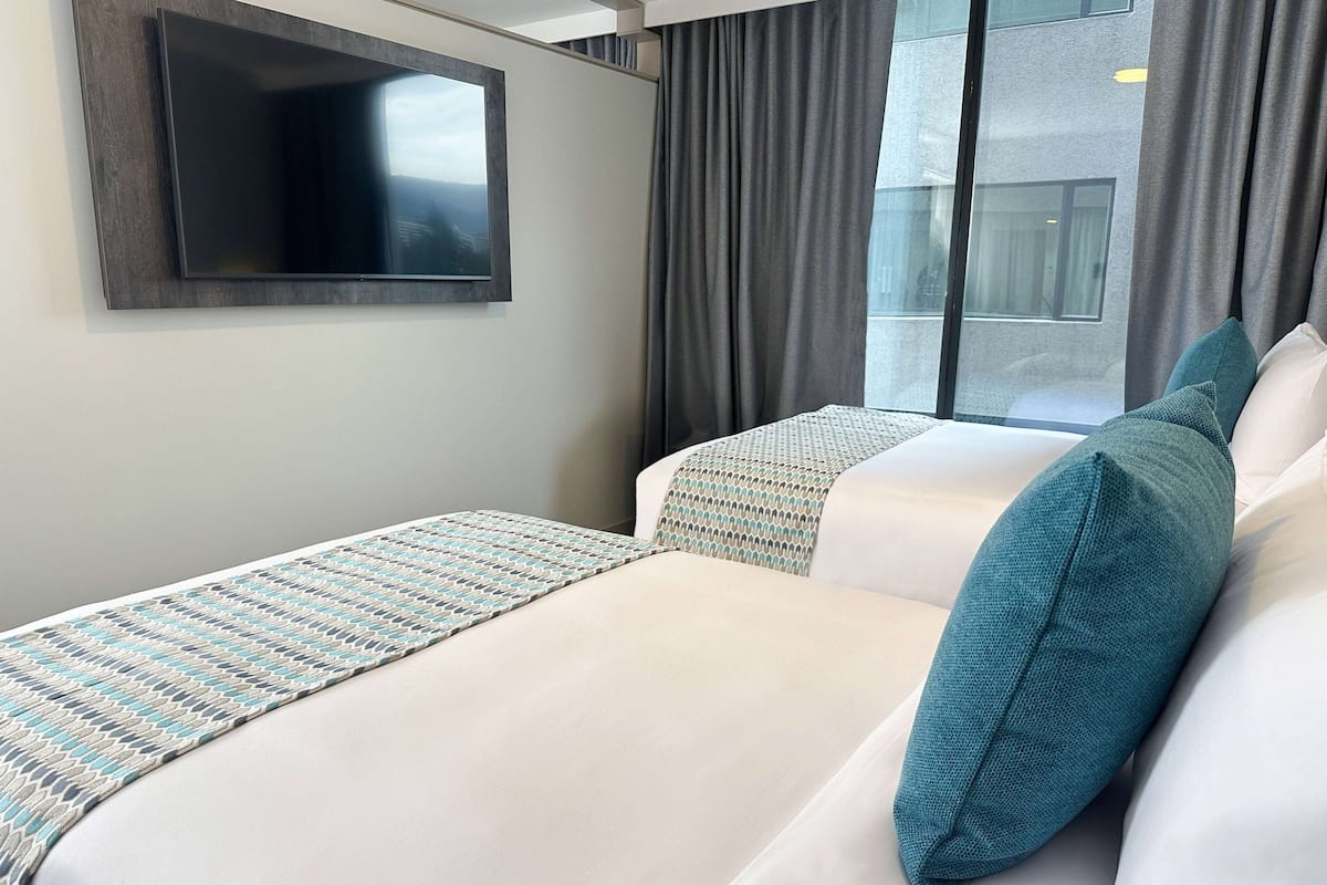 A hotel room with a large flat-screen TV mounted on the wall, a bed with white bedding and teal pillows, and a window with grey curtains.
