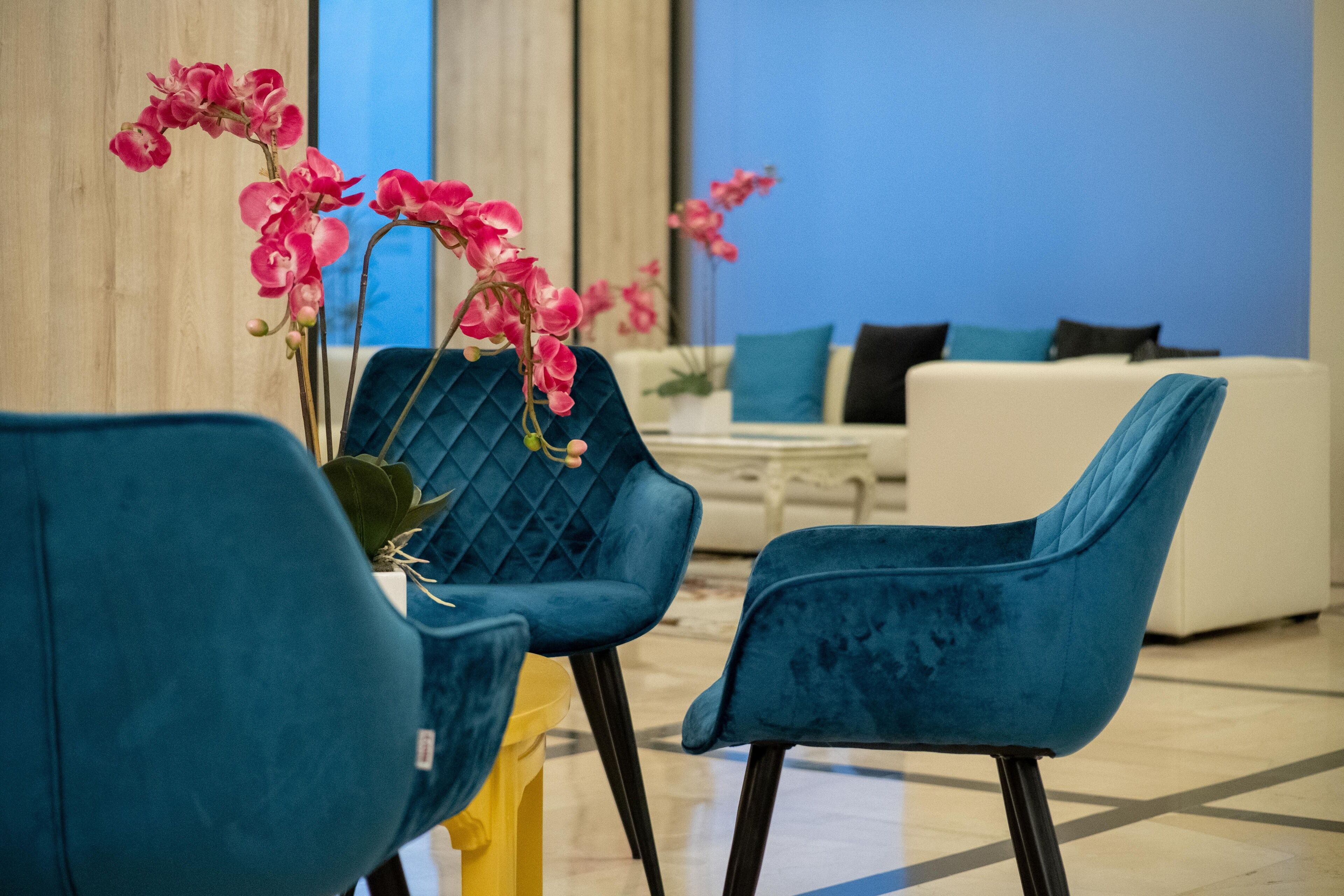 Una sala de estar moderna con sillas tapizadas en color verde azulado, un sofá blanco con cojines azules y negros, y orquídeas rosadas en un jarrón.