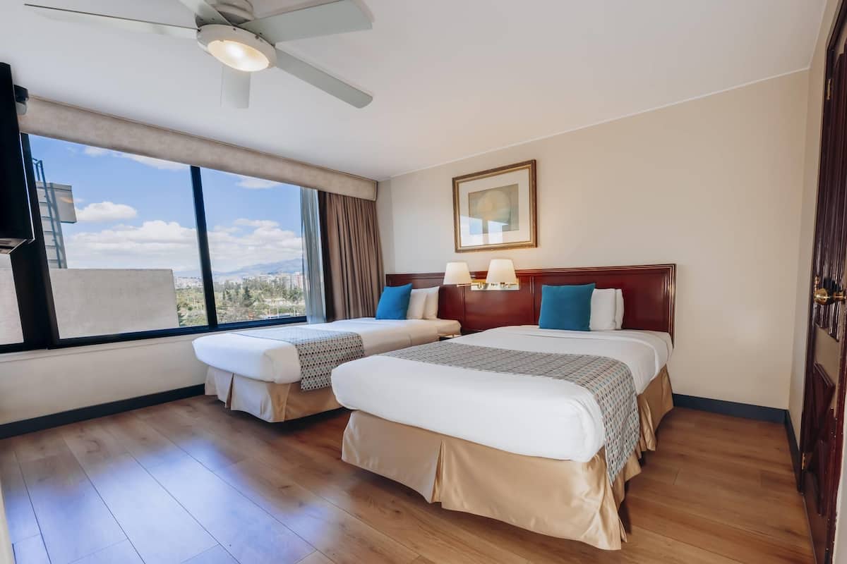 A hotel room with two beds, a ceiling fan, a large window with a view, and a framed picture on the wall.
