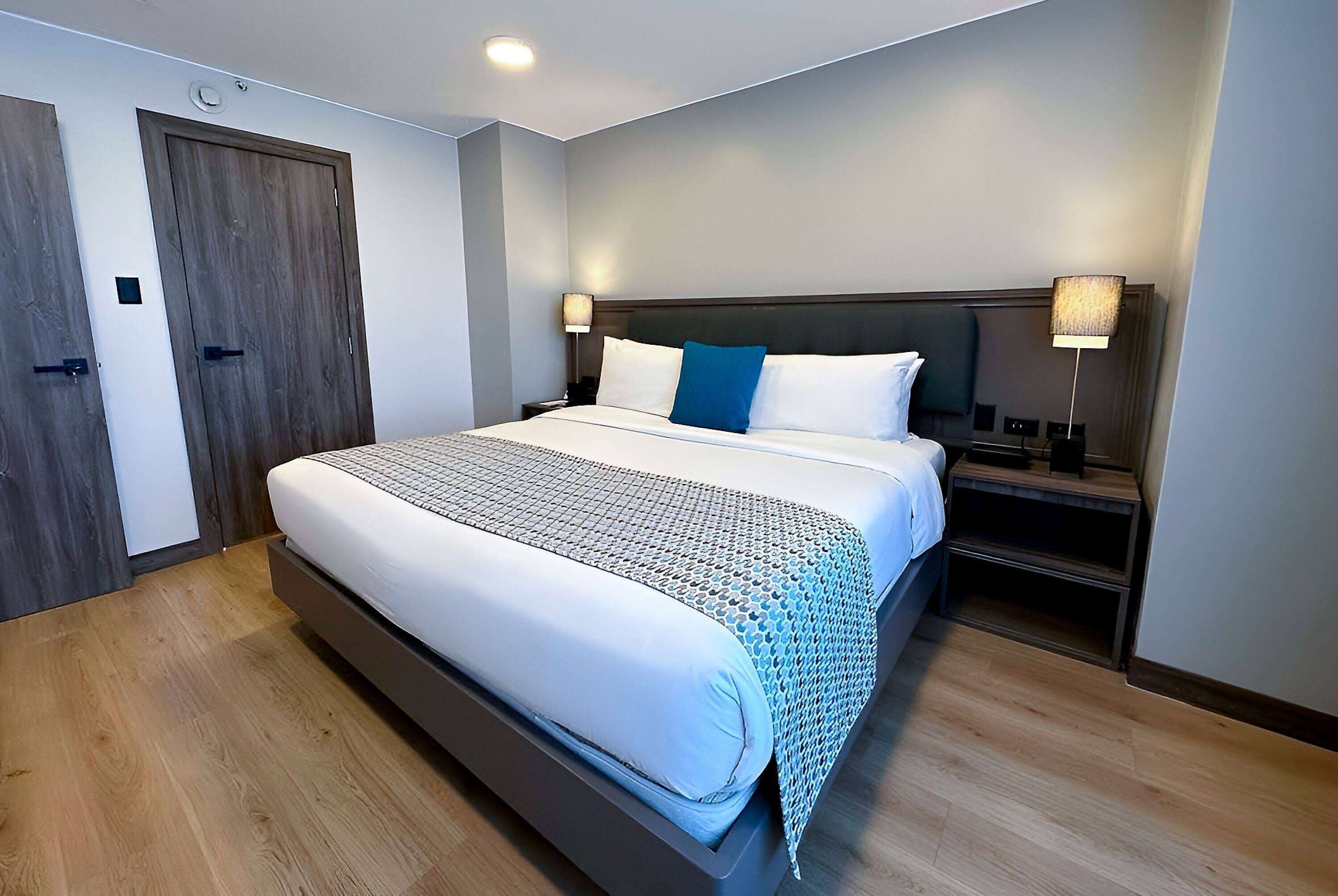 A modern hotel room with a large bed, wooden flooring, and two bedside tables with lamps.
