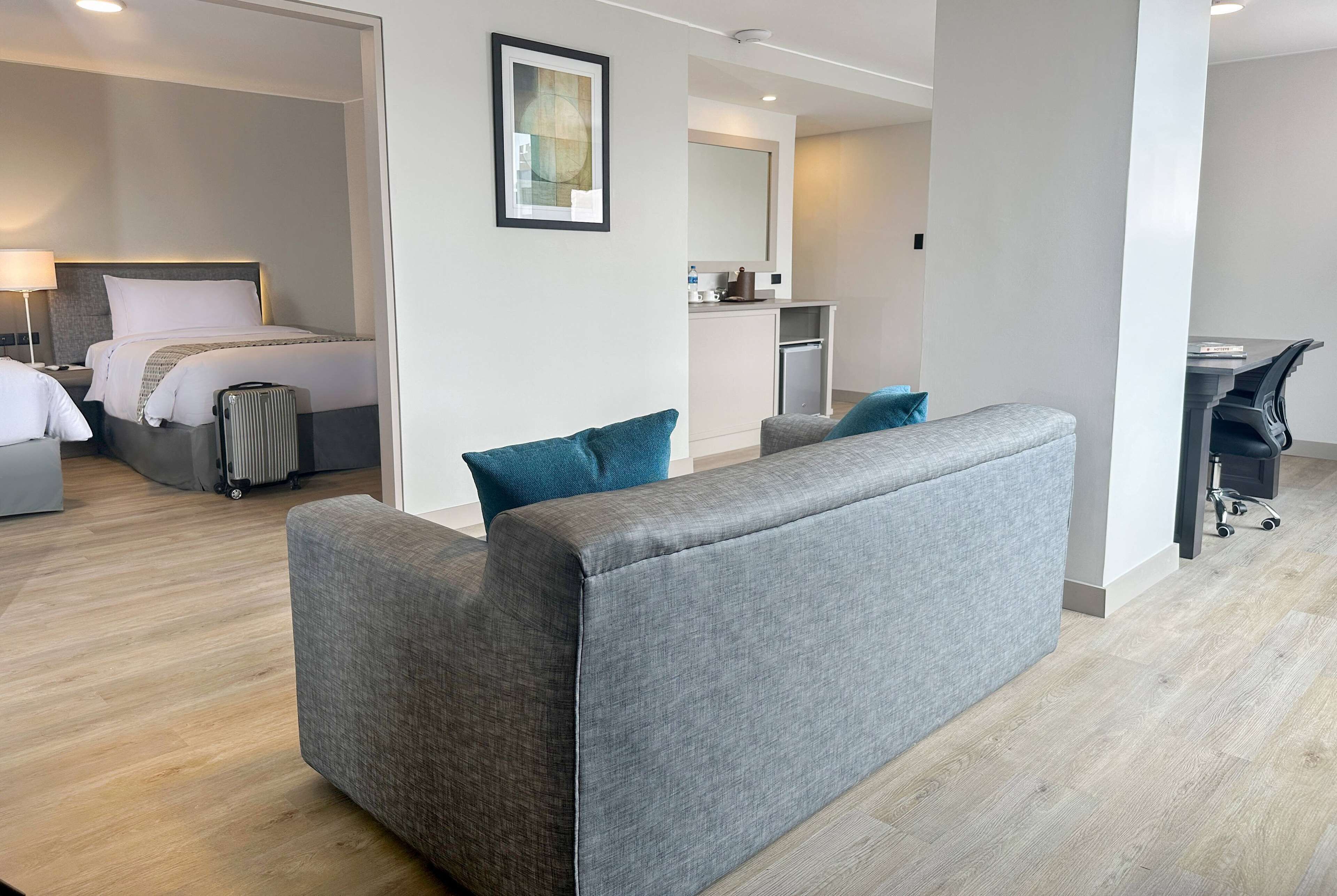 Una habitación de hotel moderna con un sofá gris, una cama con ropa blanca, una kitchenette pequeña y un escritorio con una silla.