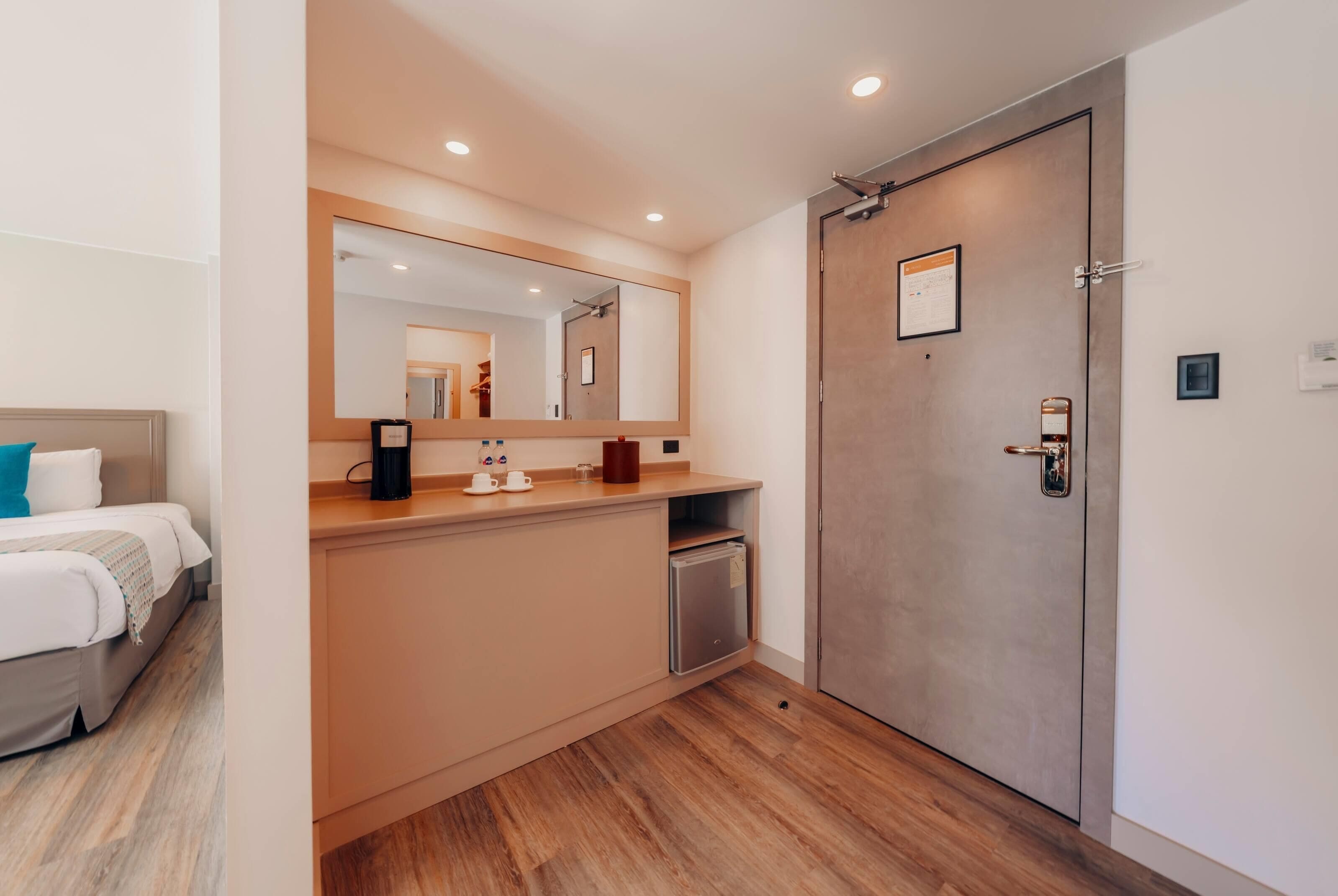 A hotel room with a bed, a bedside table with a coffee maker, a mirror, a door, and a small refrigerator.