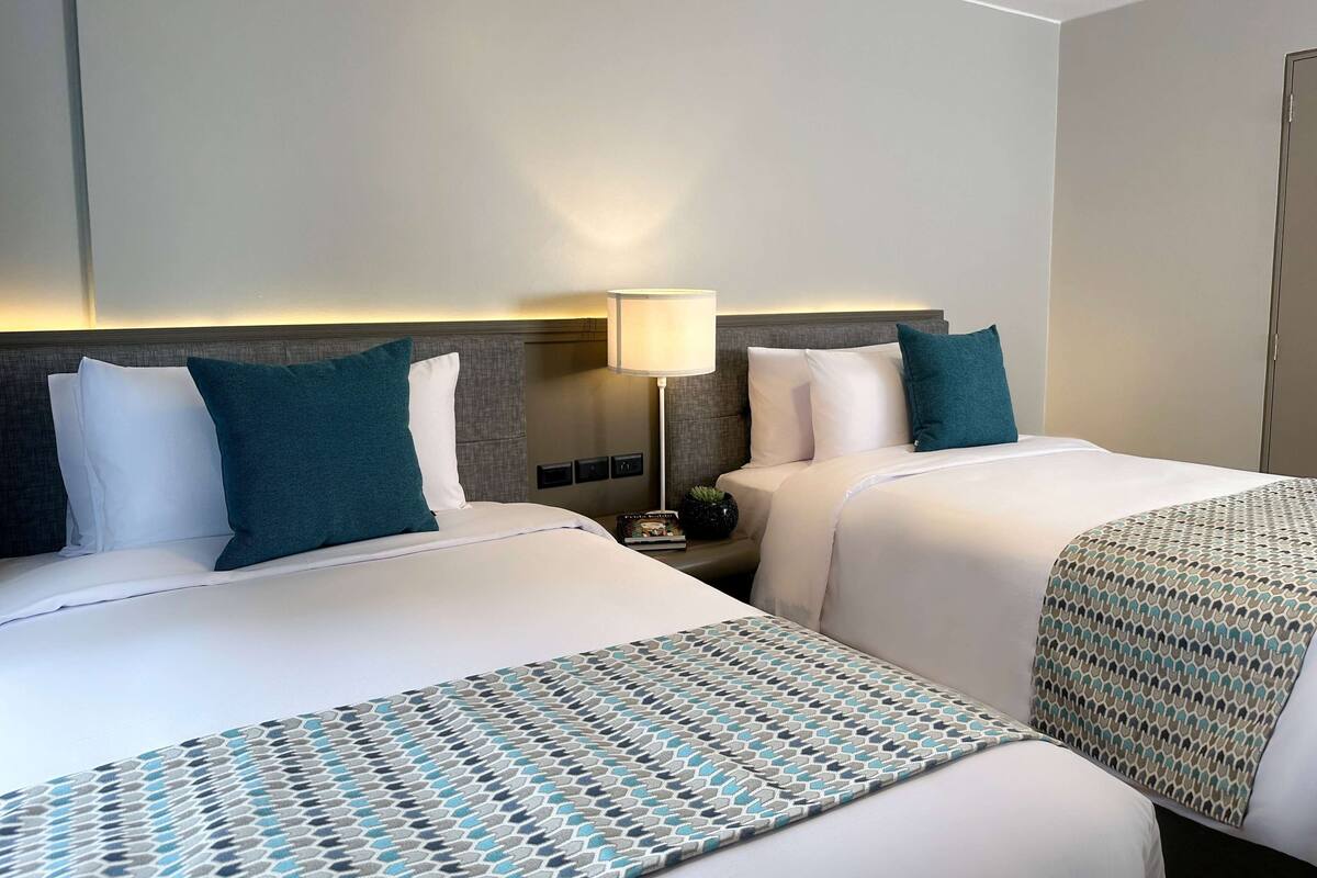 A hotel room with two beds, a nightstand, a lamp, and a patterned throw blanket.
