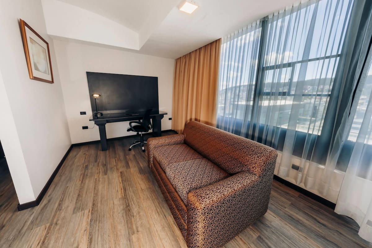 A modern hotel room with a sofa, a desk with a computer, a TV, and large windows with curtains.