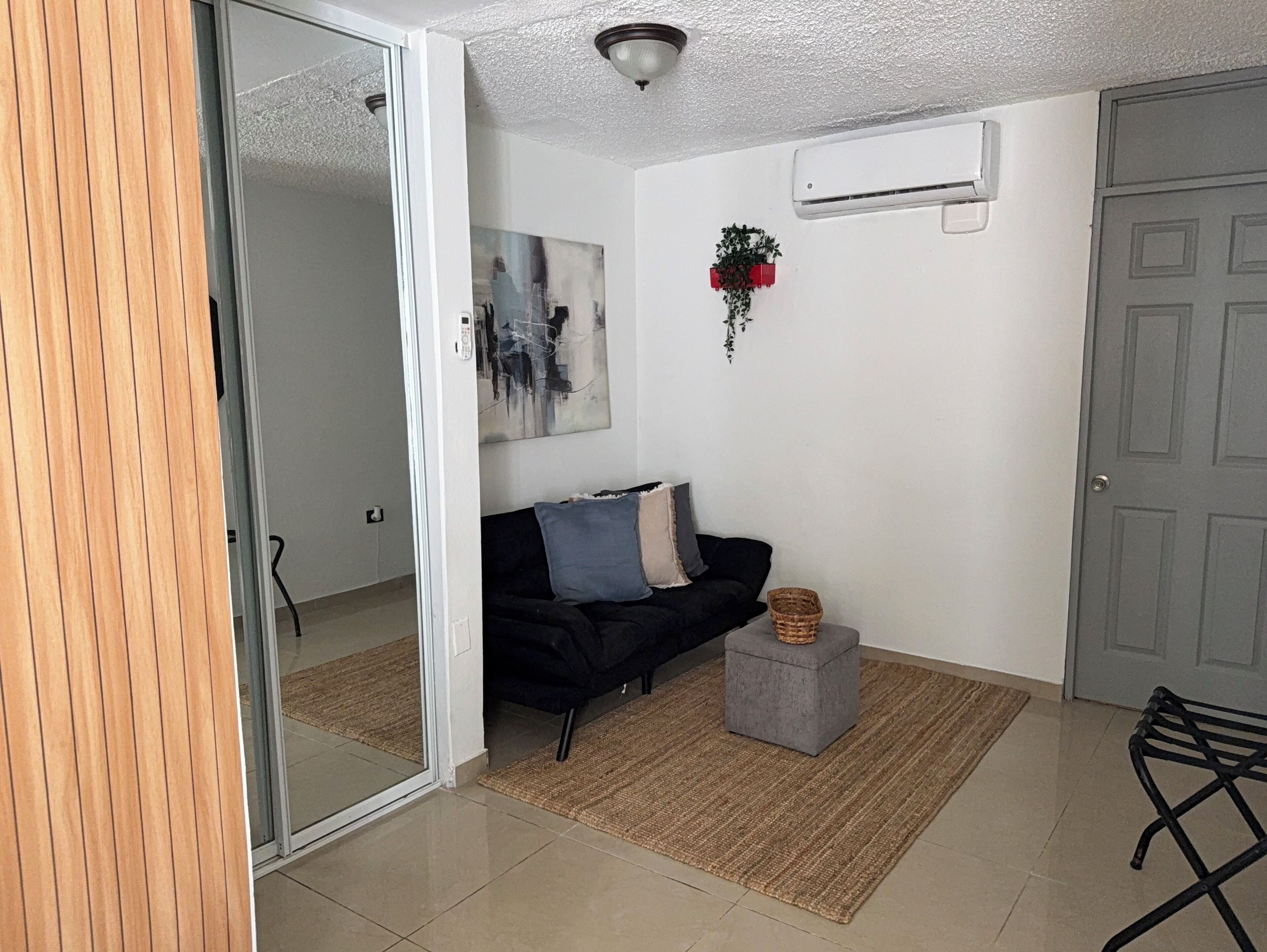 Un salon avec un canapé noir, un pouf gris, un tapis tissé et une porte grise.