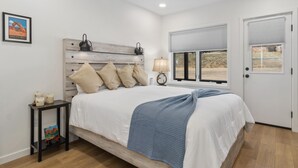 Une chambre à coucher avec un lit, une table de chevet, une lampe, un tableau encadré et une fenêtre avec des stores.