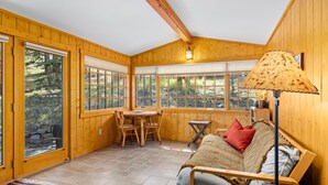 A cozy room with wooden walls, a bed, a sofa, a table, and large windows offering a view of the outdoors.