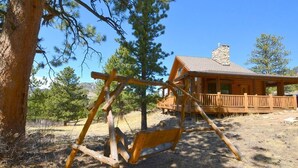 Ein rustikales Holzschaukelgestell in einer trockenen, grasbewachsenen Gegend, im Hintergrund eine Holzhütte.
