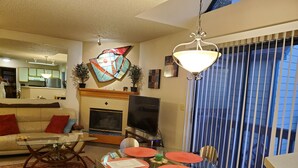 Una sala de estar con sofá, televisión, chimenea, mesa de comedor con sillas y un gran ventanal con persianas.