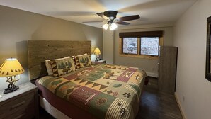 Quarto com cabeceira de madeira, colcha acolchoada, ventilador de teto e janela com vista para uma paisagem nevada.