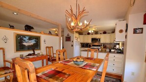 A dining area with a wooden table, chairs, and a chandelier. There is a kitchen in the background with white cabinets and appliances. - Twin Sisters - 2 bedroom 2 bathroom townhouse (Estes Park)
