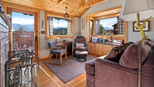 Ein gemütliches Zimmer mit Holzbalkendecke, einer Couch, einem Sessel, einem Schreibtisch und Blick auf Berge durch große Fenster.