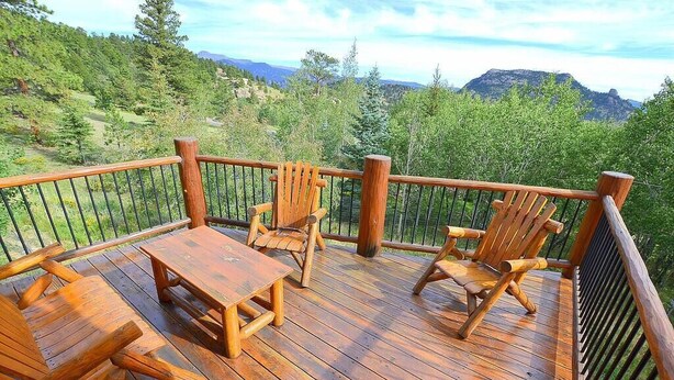 Una terraza de madera con mesa y sillas, rodeada por una barandilla metálica, con vistas a un paisaje montañoso.