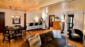 A spacious living area with a wooden ceiling, a dining table with chairs, a sofa with cushions, and a staircase leading to another level.