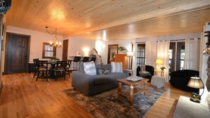 A spacious living room with a wooden ceiling, a grey sofa, a dining table with chairs, and a black armchair.