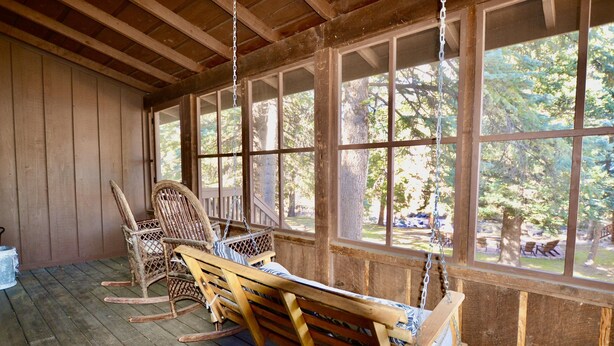 A wooden porch with rocking chairs and a swing, large windows, and a view of trees.