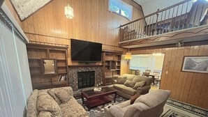 A spacious living room with a fireplace, a television mounted above it, and a balcony with a railing. - Lodgepole - 2 bedroom monthly rental - Free WiFi (Estes Park)