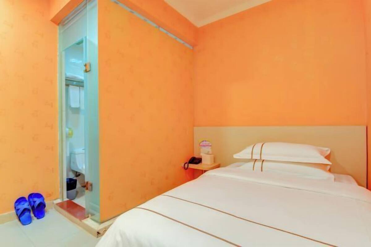 Habitación de hotel con paredes naranja, una cama con ropa blanca, una mesita de noche con teléfono y una cabina de ducha de vidrio.