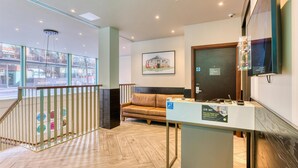 A reception area with a reception desk, a sofa, a framed picture on the wall, and a view of the outside through large windows. - Hub By Premier Inn London Camden (London)