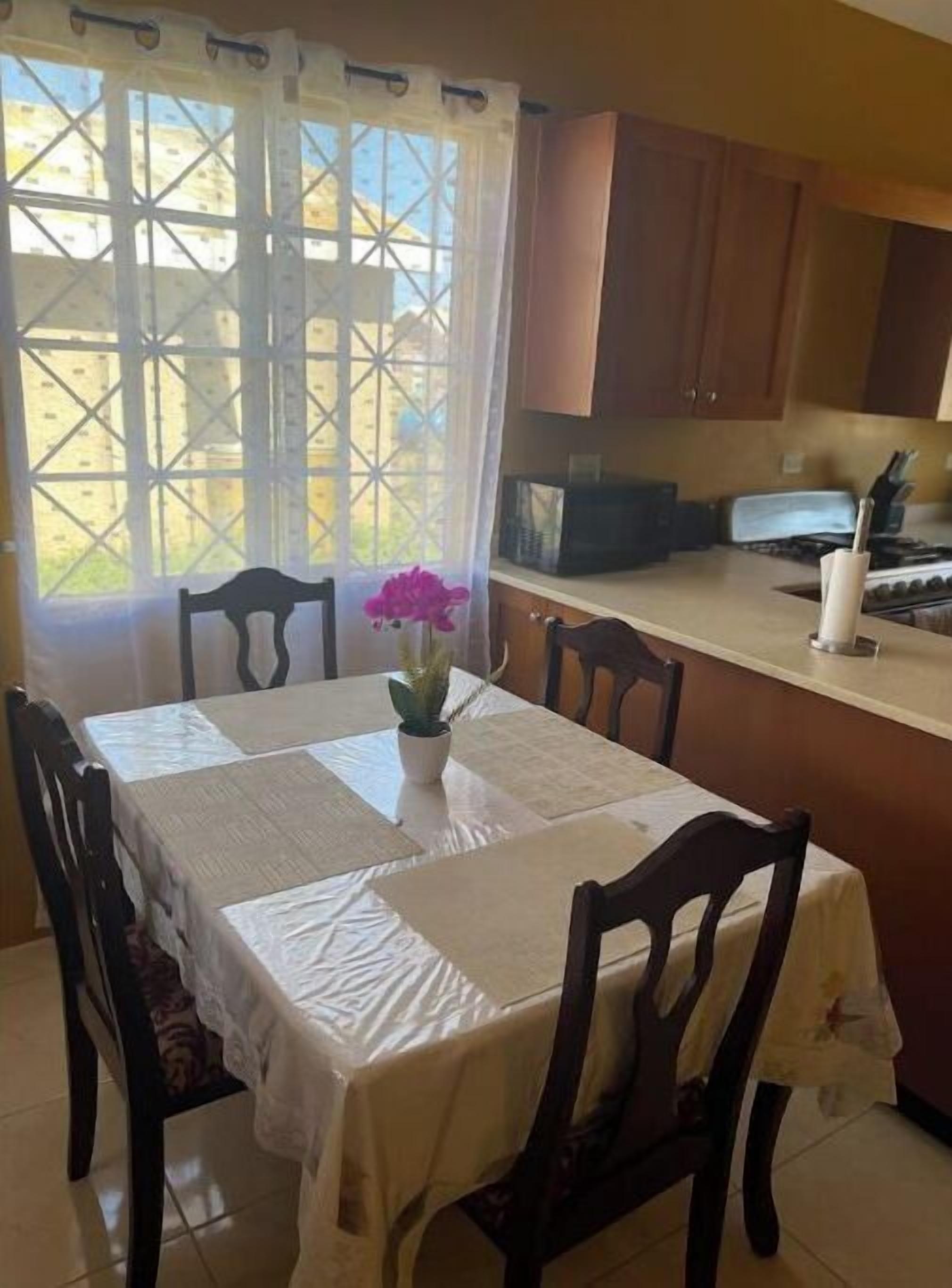 A dining area with a table covered in a white tablecloth, surrounded by wooden chairs. A potted plant with purple flowers is on the table.