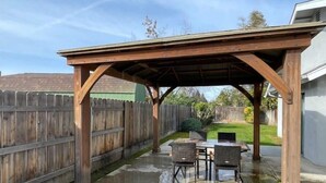 En overdækket terrasse med et trætag, udendørs møbler og et træhegn i baggrunden.