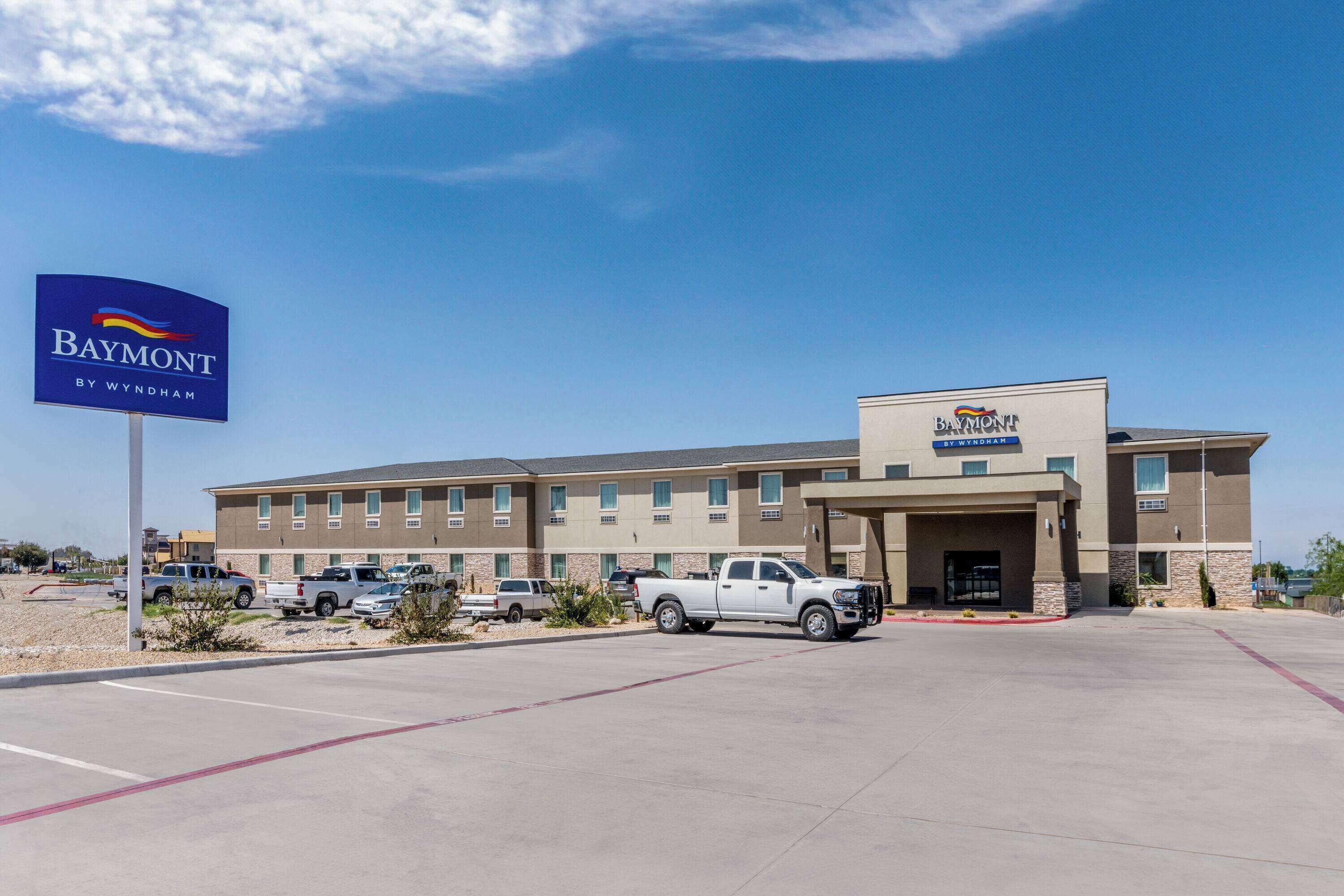 A Baymont by Wyndham hotel with a parking lot, several parked vehicles, and a clear blue sky.