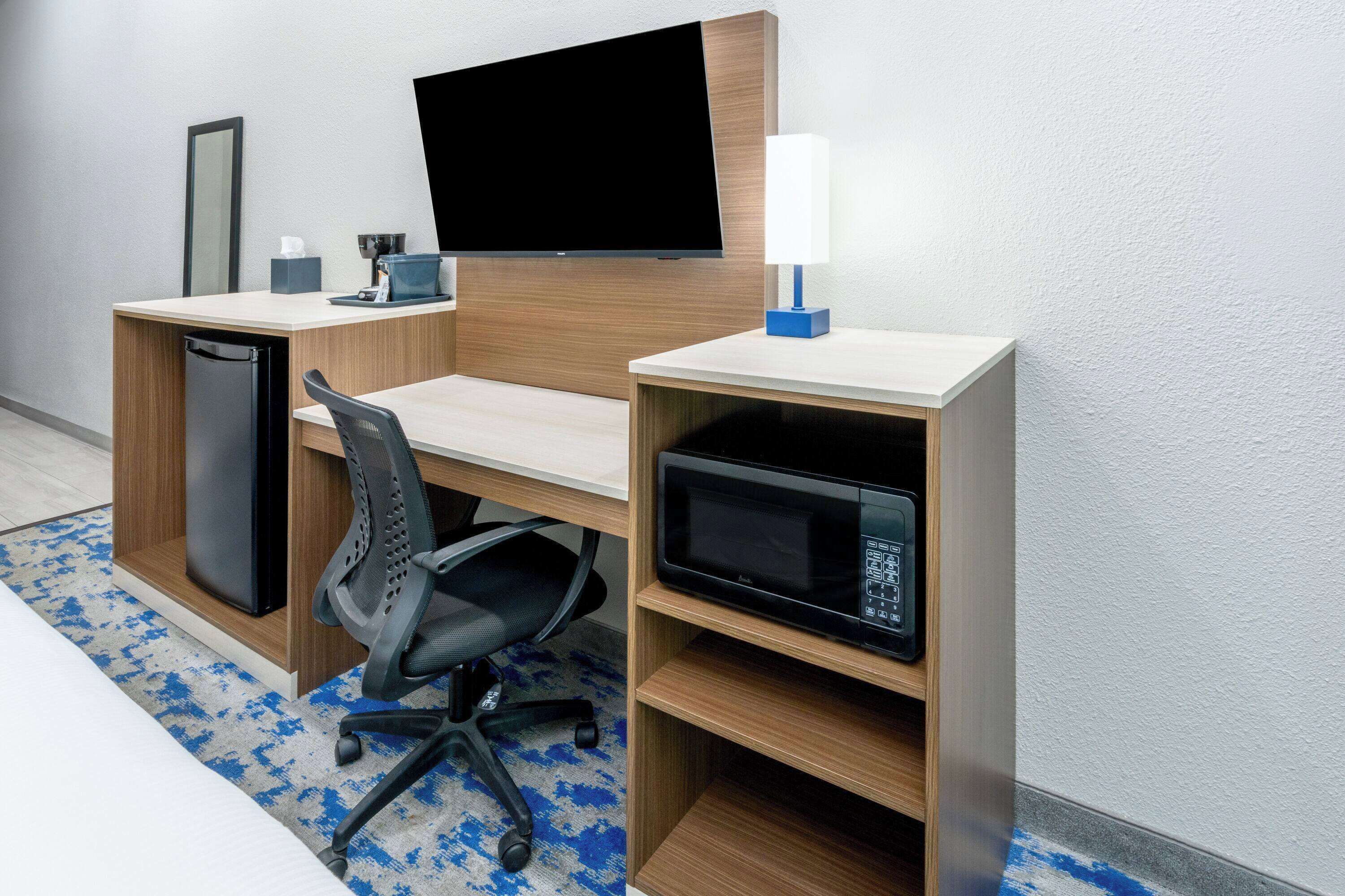 A hotel room with a wooden desk, a flat-screen TV, a microwave, a coffee maker, a lamp, and a compact refrigerator.