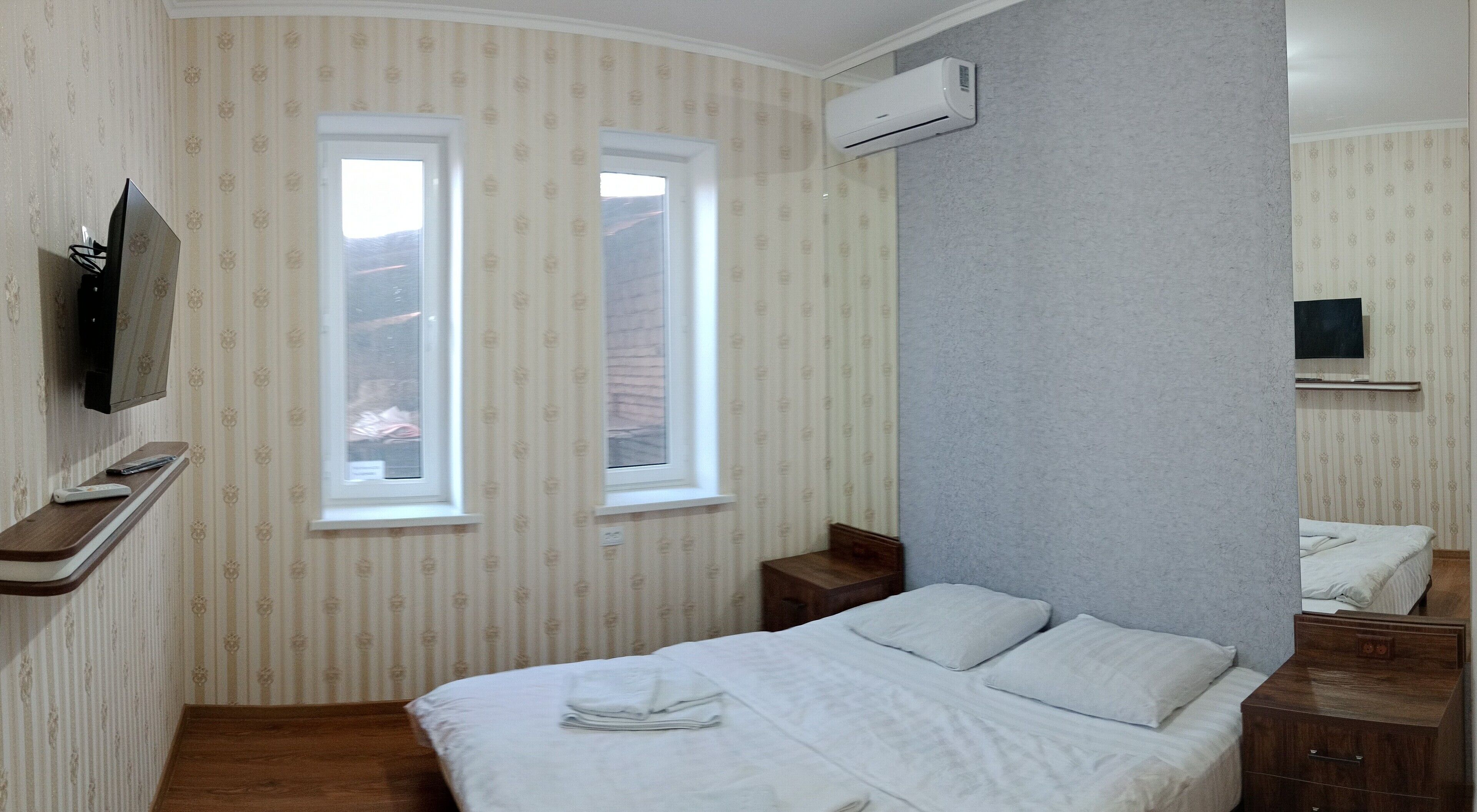 Un dormitorio con cama, dos ventanas, aire acondicionado, televisor y mesita de noche de madera.