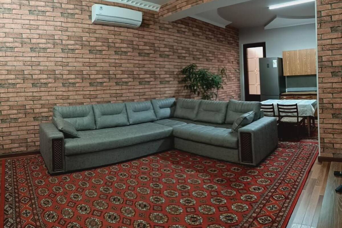 A living room with a grey sectional sofa, a red patterned rug, a brick wall, and a kitchen area in the background.