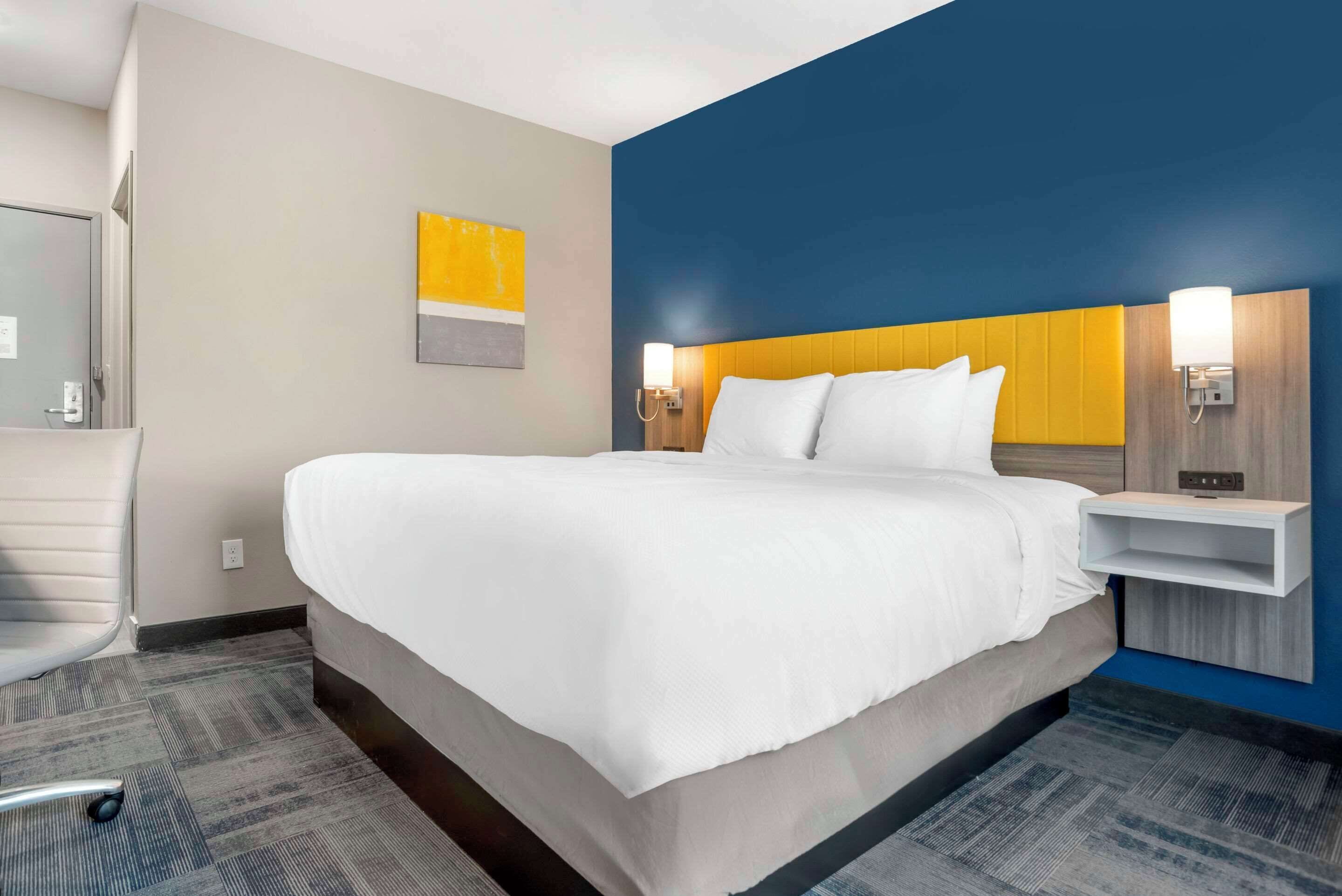 Une chambre d’hôtel avec un grand lit, une chaise, une étagère fixée au mur et un tableau.