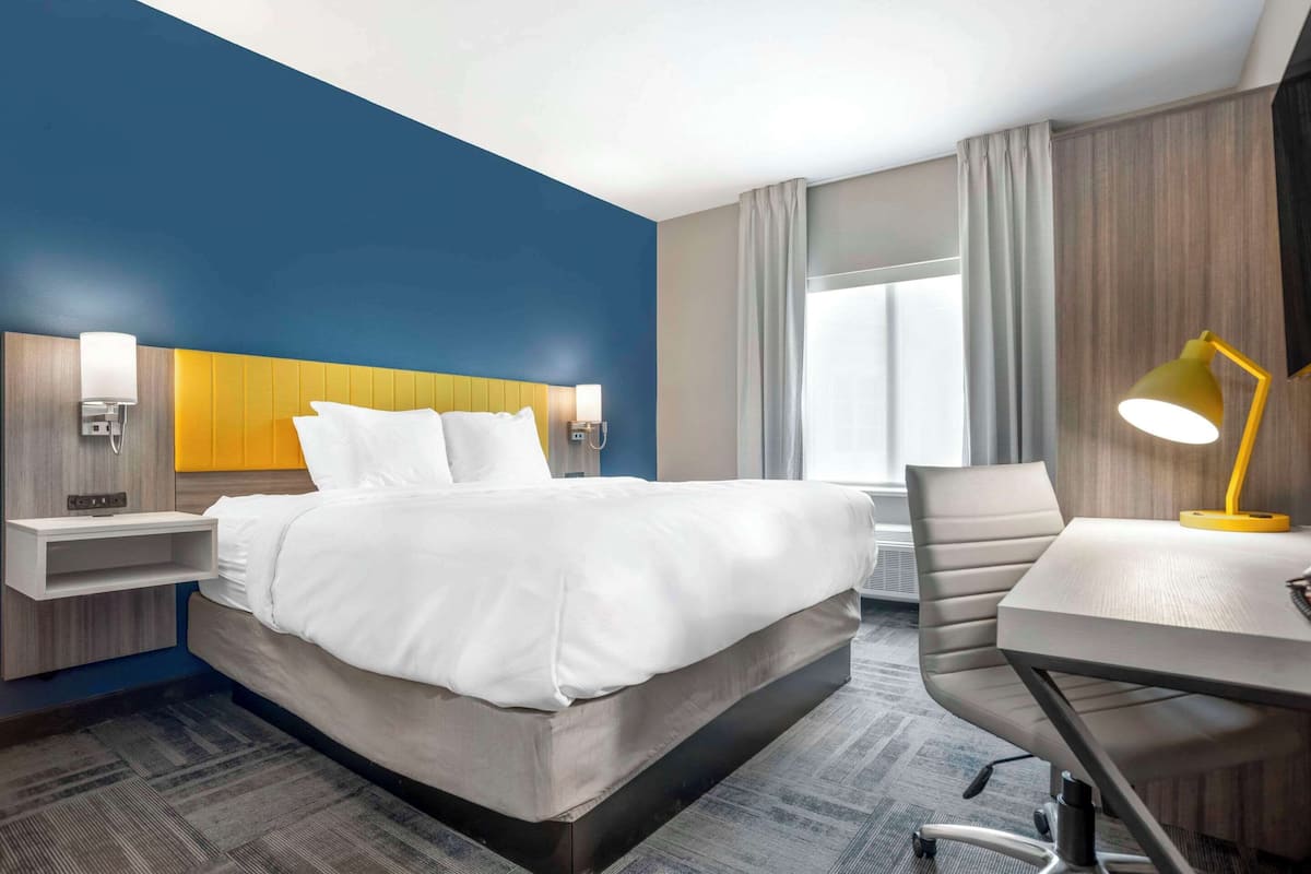 A hotel room with a bed, bedside tables, a desk with a chair, and a large window with curtains.