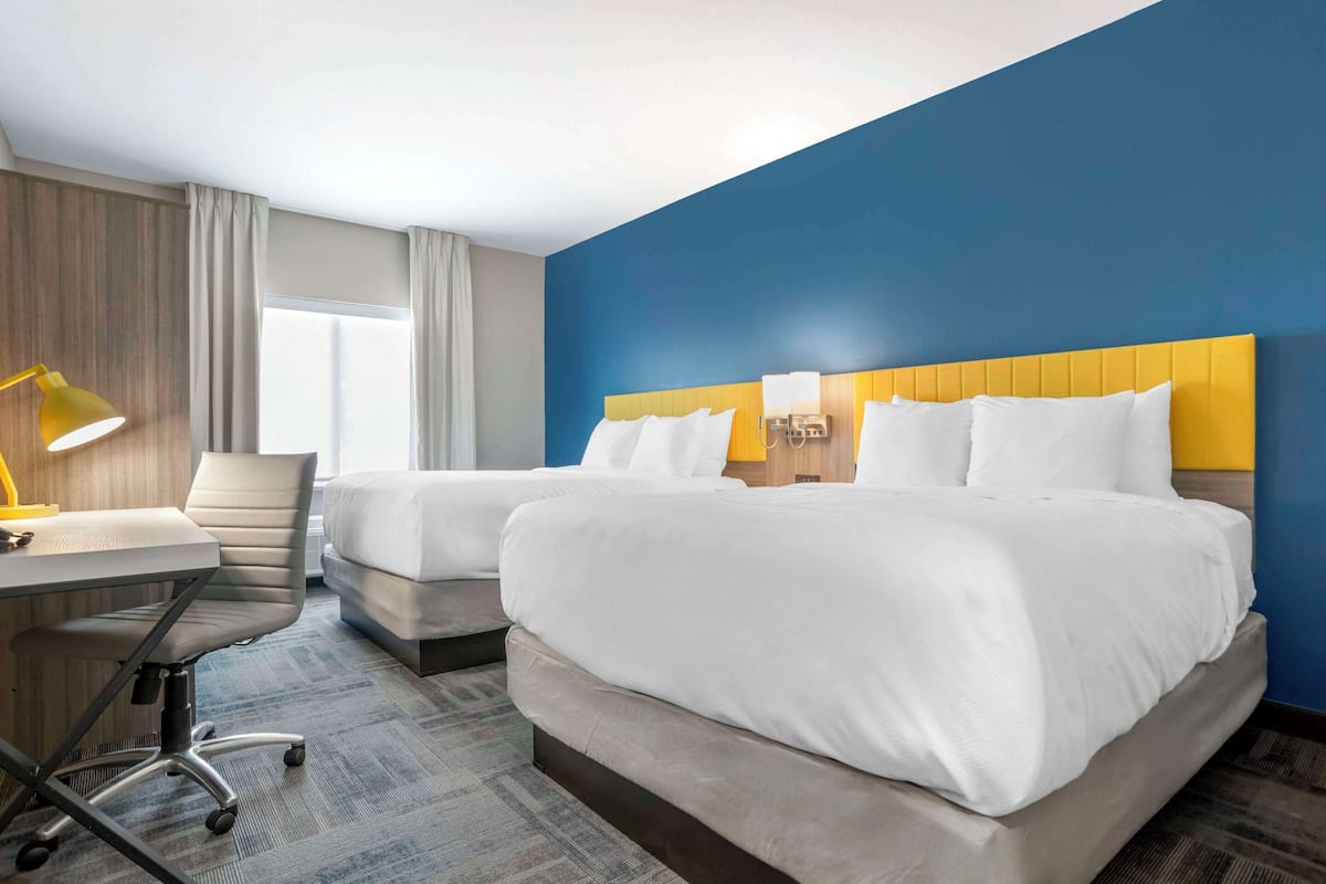 A hotel room with two beds, a desk with a lamp, and a view of the outdoors.