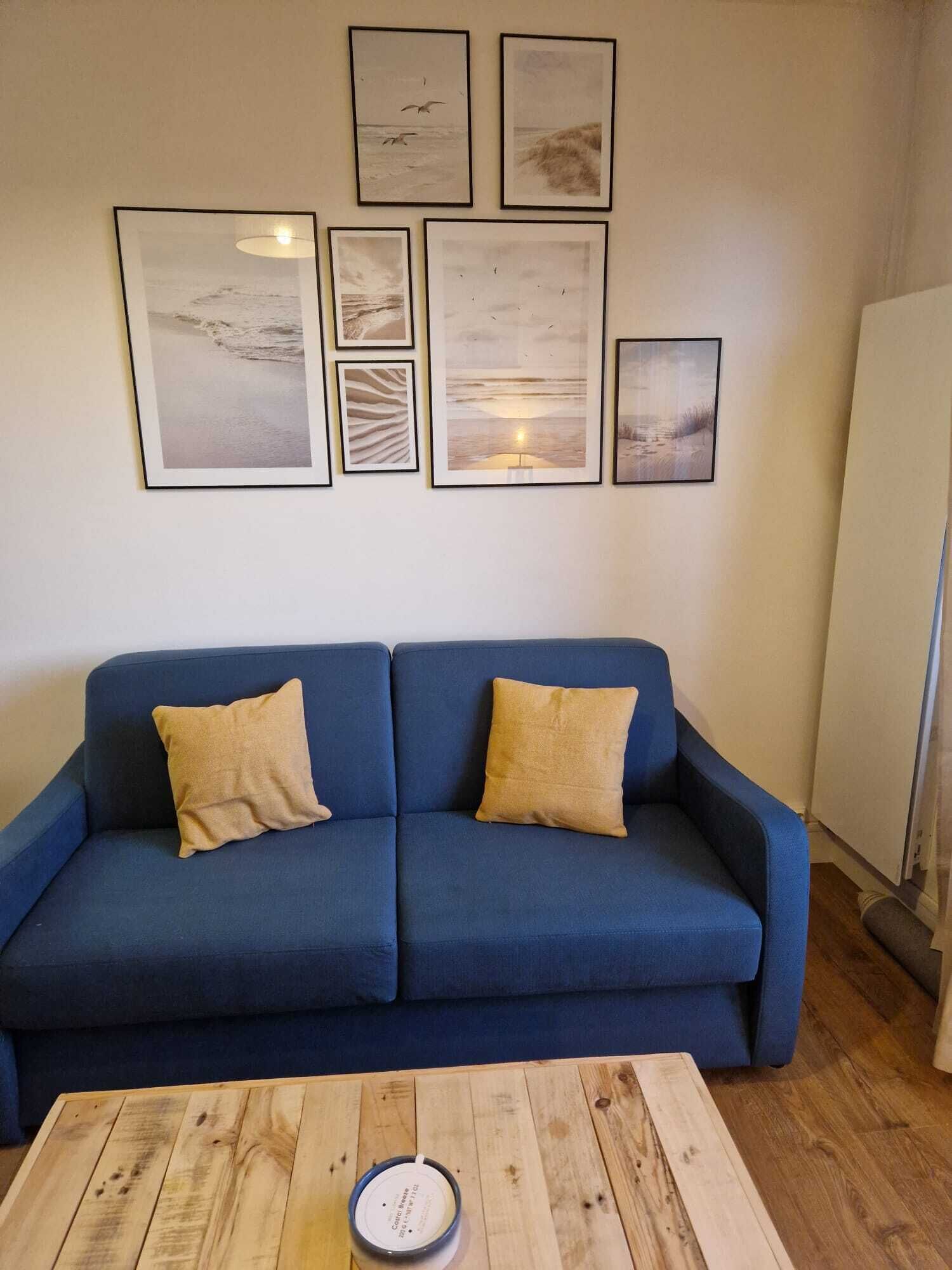 En blå sofa med to beige puder, et træbord og en væg med seks billeder i rammer.