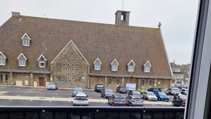 A balcony with a black metal table and a wooden bench, overlooking a parking area with several cars and a building with a distinctive roof and multiple windows. - Le Portel Plage (Le Portel)