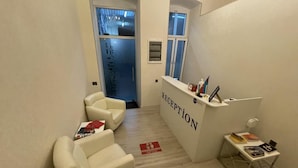 A reception area with a reception desk, two white armchairs, a small table with books, and a window with a view of the outside. - Solo Hotel  (Baku)