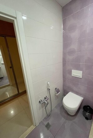 Une salle de bain avec des murs carrelés de blanc et de violet, des toilettes blanches, une douche et une poubelle noire.