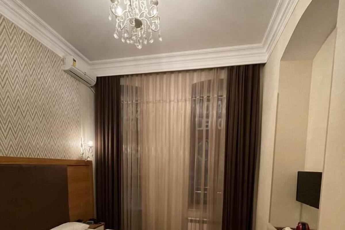 A hotel room with two beds, a chandelier, a desk with a chair, and a large window with curtains.