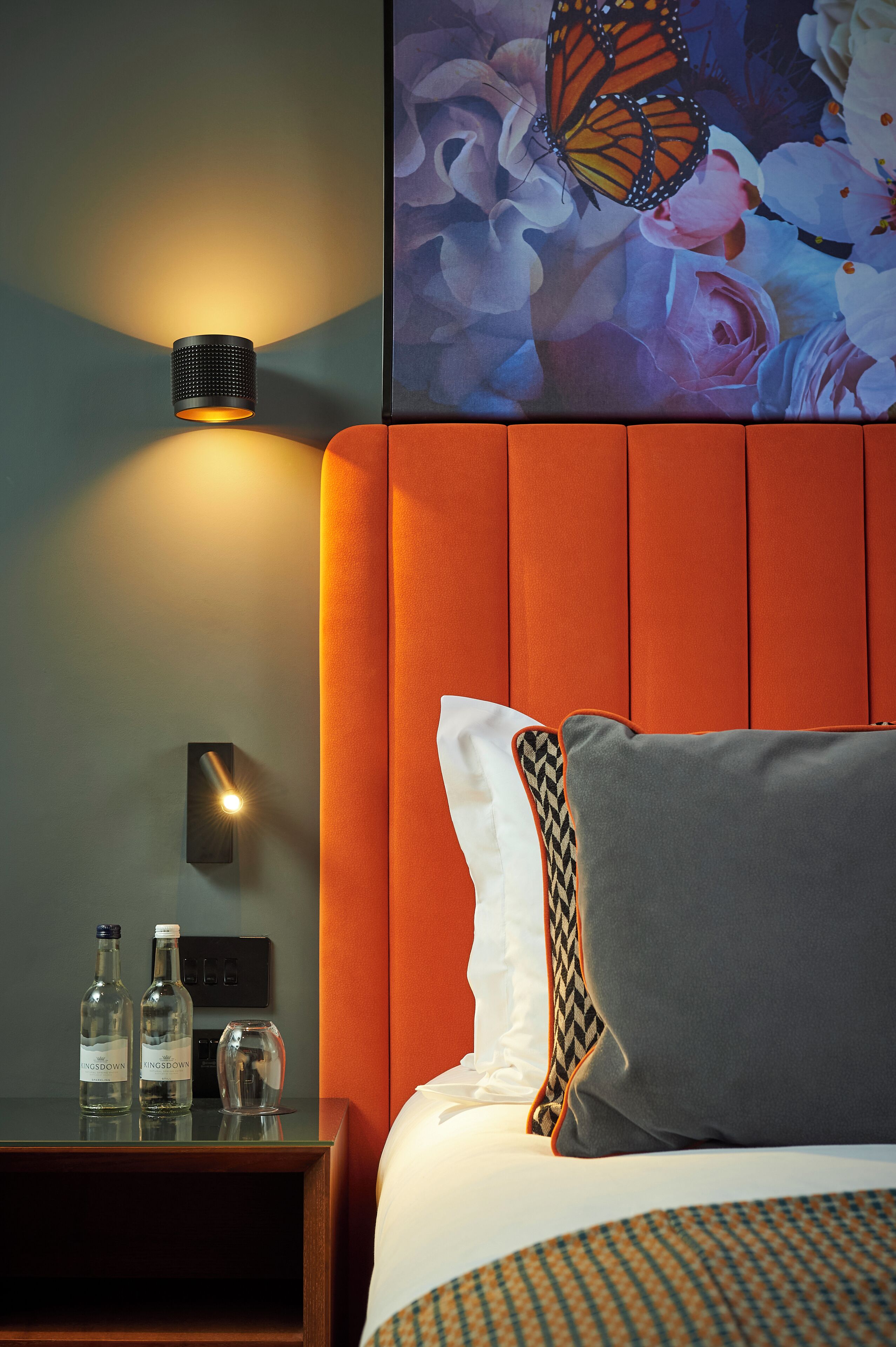 Habitación de hotel con una cama, una mesita de noche con botellas, una lámpara de pared y un mural de mariposas.