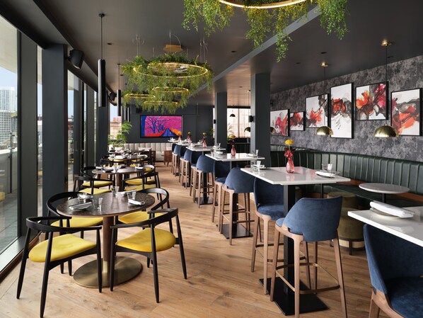 A modern restaurant interior with wooden flooring, blue and yellow chairs, and a large window offering a city view.