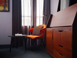 Una habitación con un armario de madera, una mesa redonda con un libro y vasos, una silla naranja con un cojín estampado, y una ventana con cortinas.