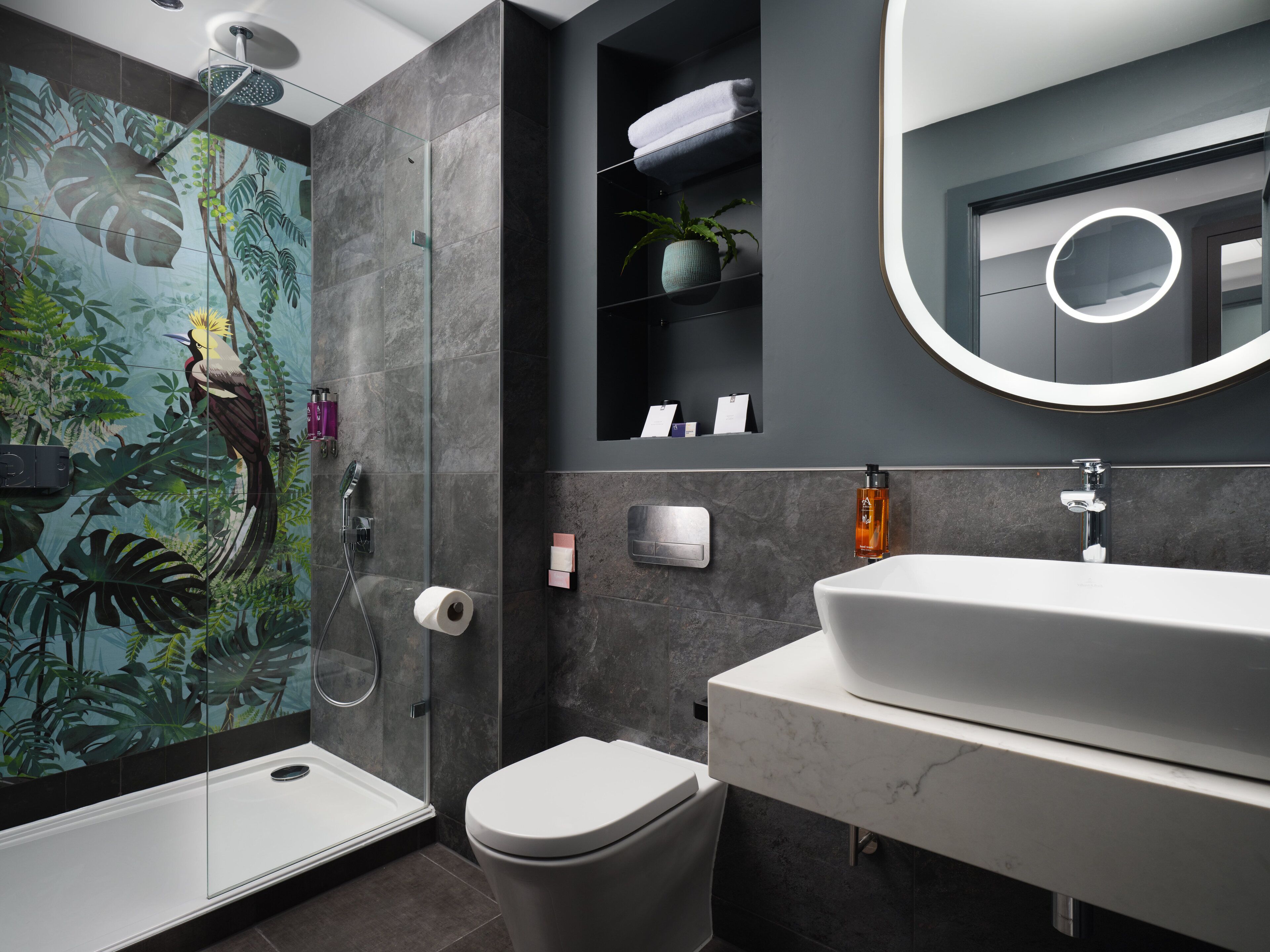 Un baño moderno con ducha de vidrio, inodoro, lavamanos con encimera de mármol, espejo con marco circular y estante con toallas y planta en maceta.