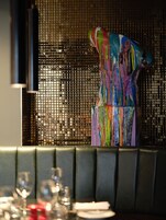 Un comedor con una escultura abstracta de colores, un separador de malla negra, y una mesa dispuesta con vasos y platos.