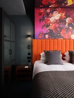 Habitación de hotel con una cama grande, una mesita de noche, un aparador de vidrio y una obra de arte floral colorida en la pared.