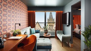 A modern living room with a sofa, armchairs, a coffee table, and a desk with a laptop. A large window offers a view of a building with a pointed roof. - Malmaison Manchester Deansgate  (Manchester)