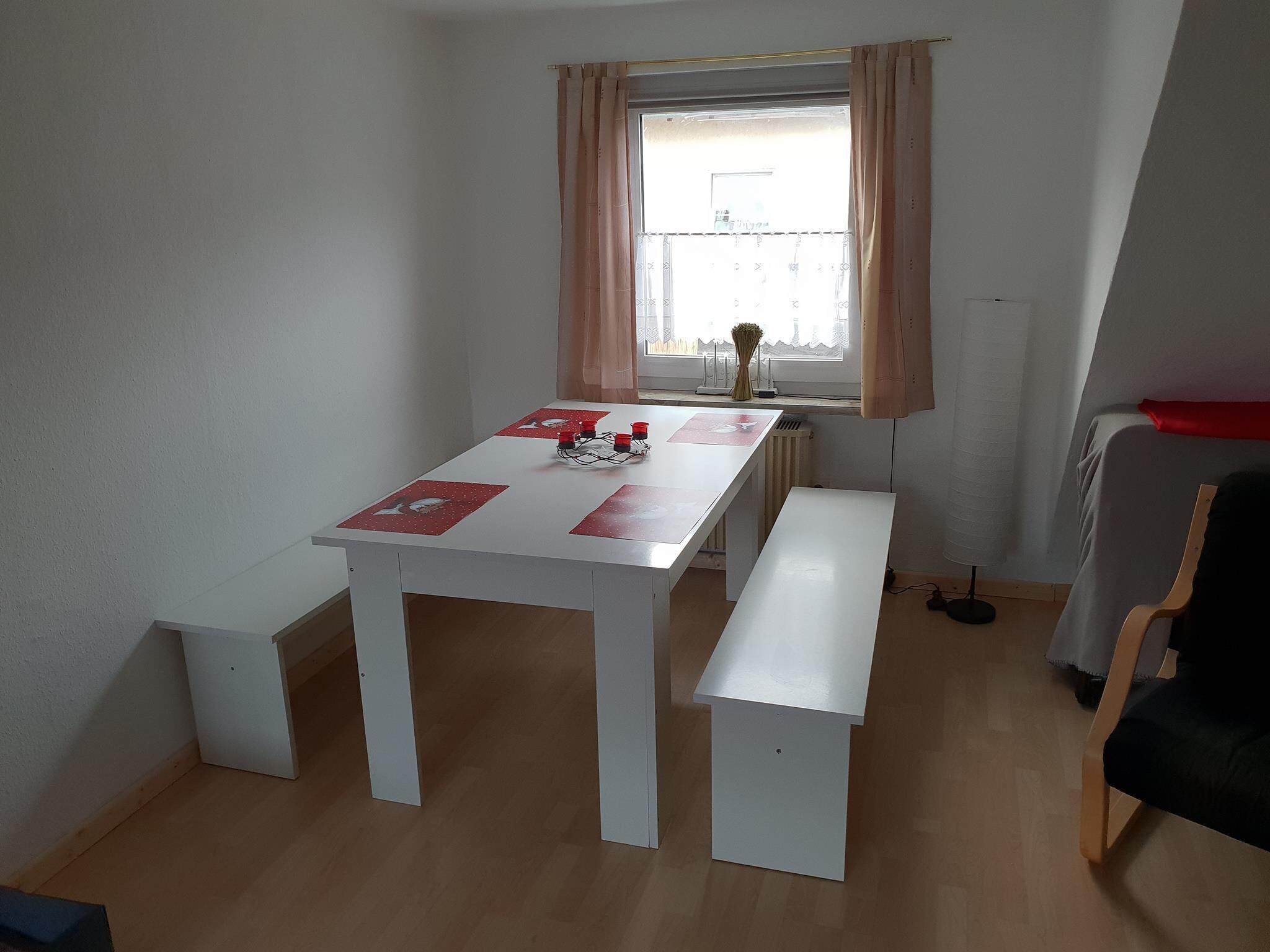 Une petite pièce bien éclairée, avec une table et des bancs blancs, une fenêtre avec des rideaux et un radiateur.