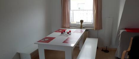 Une petite pièce bien éclairée, avec une table et des bancs blancs, une fenêtre avec des rideaux et un radiateur.