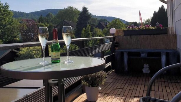 Una terraza con una mesa de vidrio, dos copas de vino blanco y una botella de vino. Al fondo, se observa un paisaje montañoso.