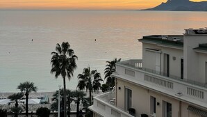 A coastal view with a mountain in the distance, palm trees, and a building with balconies. - Apartment with Partial Sea View – Sunset – Cannes Croisette – Palais Miramar (Cannes)