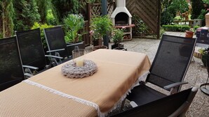 An outdoor patio with a table set for two, surrounded by chairs, a brick oven, and various potted plants. - Vacation apartment Ramke EG (Lilienthal)