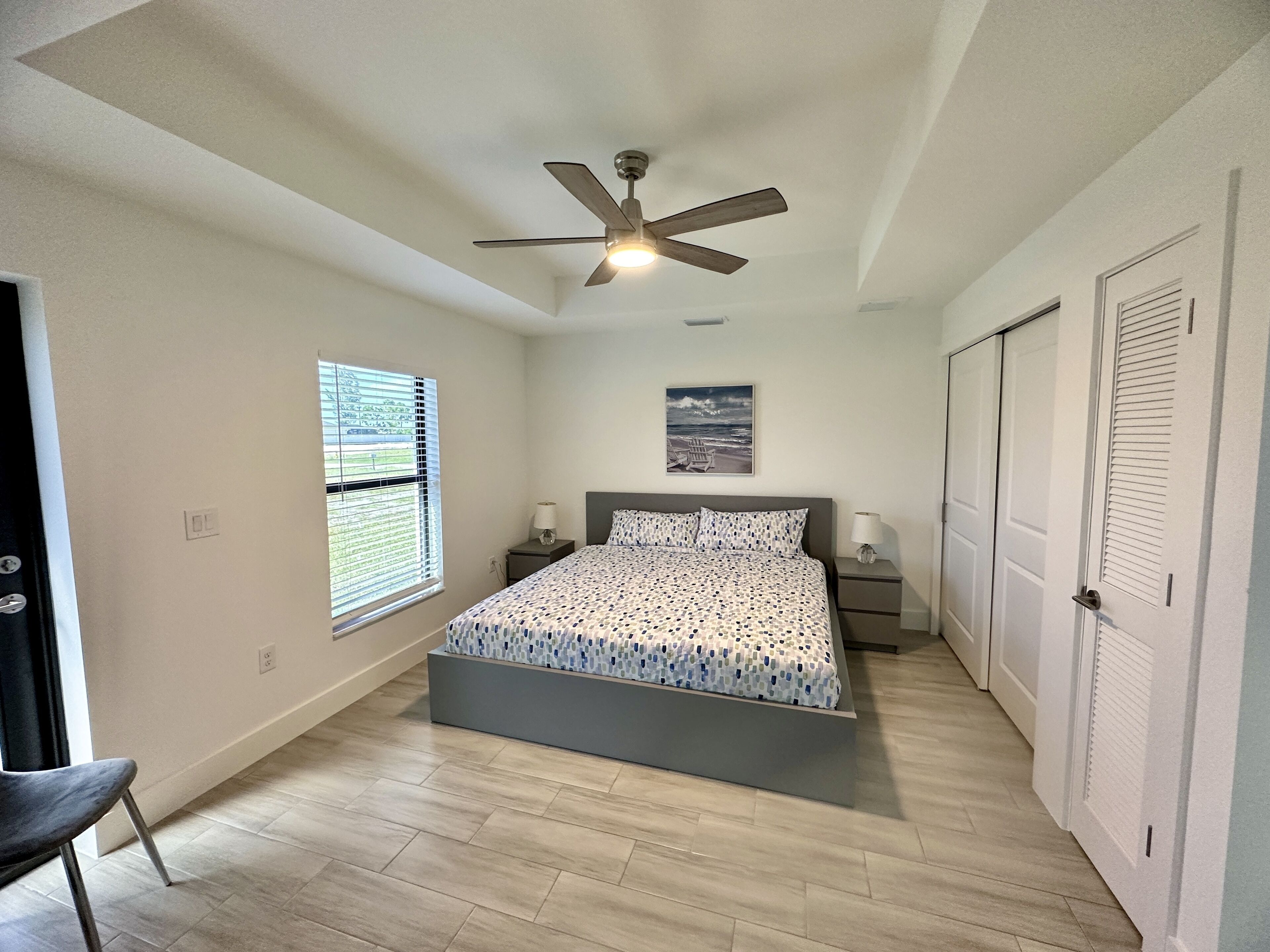 A bedroom with a bed, a ceiling fan, a window with blinds, a nightstand, a bedside table, and a door.